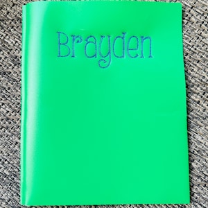 May include: A bright green notebook with the name "Brayden" embroidered in blue thread. The notebook is closed and has a smooth, glossy finish. The background is a textured, neutral-toned surface.
