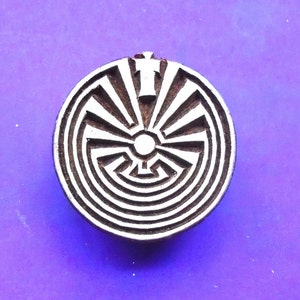 May include: A round wooden block with a carved design of a circular maze with a sunburst pattern and a cross at the top. The design is in black and white.