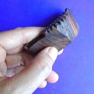 May include: A dark brown wooden comb with a textured surface. The comb has a series of teeth on one side and is held in a hand.