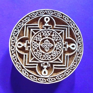 May include: A round wooden block with a carved design. The design features a square with a circle in the center, surrounded by four symbols. The block is brown and the design is white.