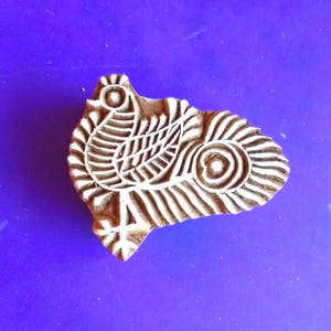 Puede incluir: Un sello de impresión de madera con un diseño detallado de un pájaro con un ojo grande y redondo. El sello es marrón y el diseño es blanco.