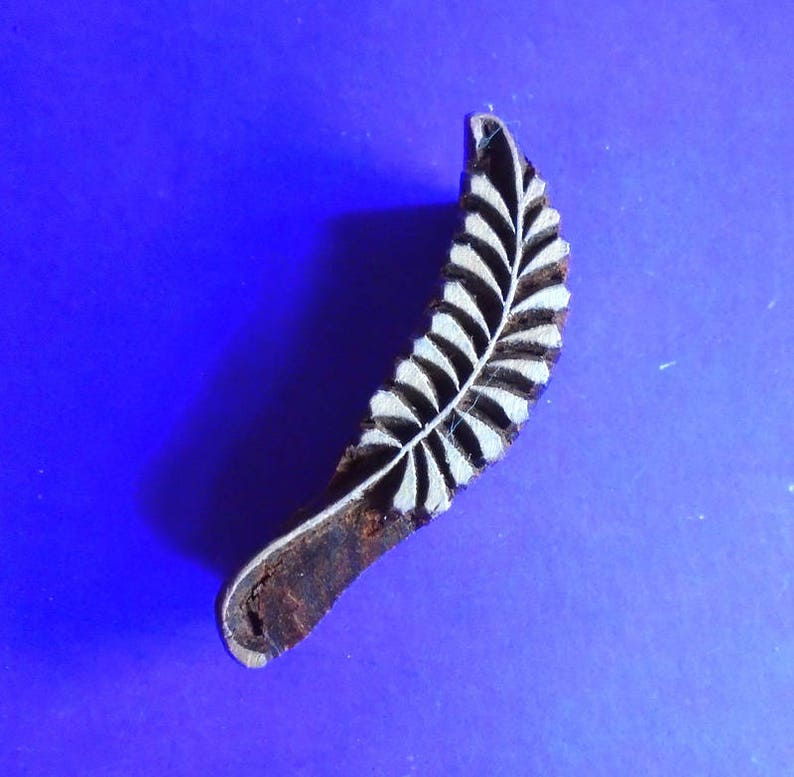 May include: A wooden block printing stamp with a carved design of a fern leaf. The stamp is brown and the leaf design is white.