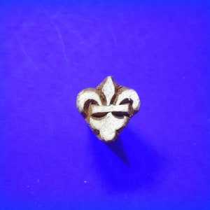 May include: A brown wooden block with a white carved design of a fleur-de-lis. The block is on a blue background.