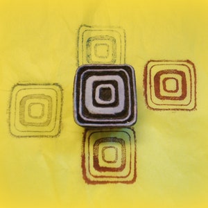 May include: A set of five black and white rubber stamps with concentric square designs. The stamps are arranged in a cross pattern on a yellow surface.