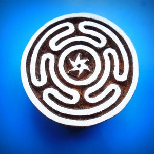 Op de afbeelding: Ronde houten stempel met een wit labyrintpatroon en een zespuntige ster in het midden. De stempel heeft een bruine achtergrond en staat tegen een blauwe achtergrond. Waarschijnlijk gebruikt voor printen of knutselen.
