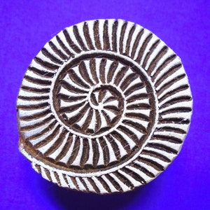 May include: A circular wooden block printing stamp featuring a carved spiral pattern. The design consists of raised white surfaces against a dark brown recessed background, creating a repeating radial motif that resembles a nautilus shell.