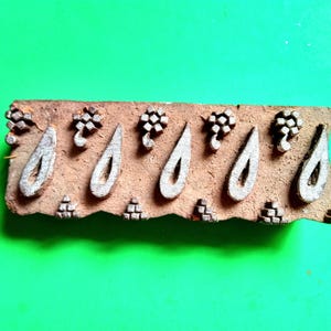 May include: A rectangular, wooden block with a repeating pattern of teardrop shapes and floral elements. The block is a light brown color with white painted designs. The background is a solid, bright green.