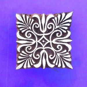 Op de afbeelding: Een vierkante houten blok met een gesneden ontwerp. Het ontwerp is een symmetrisch patroon van spiralen en bladeren, geverfd in wit op een bruine achtergrond.