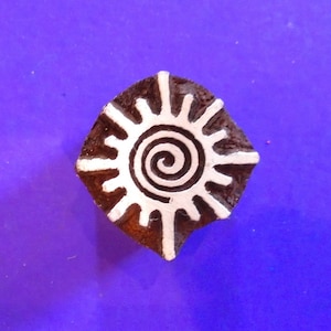 Op de afbeelding: Een houten blokstempel met een wit zonontwerp met een spiraal in het midden. De stempel bevindt zich op een paarse achtergrond.
