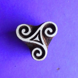 May include: A wooden block printing stamp with a three-part design of interlocking spirals. The stamp is brown with a white design and is on a purple background.