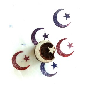 May include: A circular wooden printing block featuring a carved crescent moon and star design. The block is positioned over several stamped impressions of the same moon and star motif in red and blue ink, showing the carved relief pattern clearly.