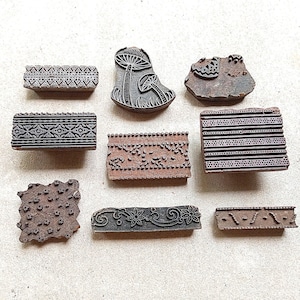 May include: A collection of eight wooden printing blocks featuring various carved designs, including geometric borders, floral patterns, and mushroom motifs. These rectangular and irregular shaped blocks have dark brown surfaces with raised relief carvings.