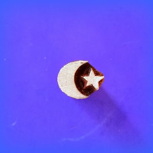 May include: A wooden block carving of a crescent moon and a five-pointed star. The carving is light brown with a darker brown outline. The background is a solid purple.