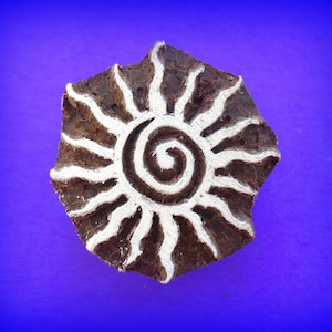 May include: A brown wooden block with a white sun design and a spiral in the center. The block is used for printing patterns on fabric.