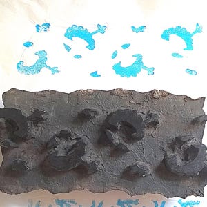 May include: A rectangular dark brown wooden block with carved raised shapes, used for printing. The block features several distinct, curved, and abstract motifs. Above the block, a series of bright cyan ink impressions show the pattern.
