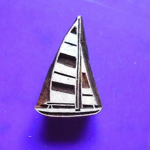 May include: A wooden block carving of a sailboat with a white sail and black lines. The carving is on a purple background.