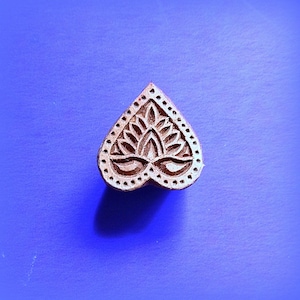 May include: A heart-shaped wooden printing block featuring a carved floral motif. The design includes a central petal pattern surrounded by a border of small dots. The natural brown wood texture contrasts with the smooth, solid blue background.