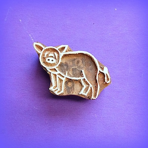 May include: A brown wooden block stamp featuring a carved outline of a pig. The stamp has a natural wood grain texture with white carved lines defining the shape of the pig, including its ears, snout, legs, and a curled tail on the right side.