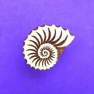 May include: A wooden printing block featuring a carved ammonite fossil design. The stamp has a light-colored, smooth surface with dark brown, recessed grooves forming the spiral shell pattern, showcasing intricate, curved lines throughout.
