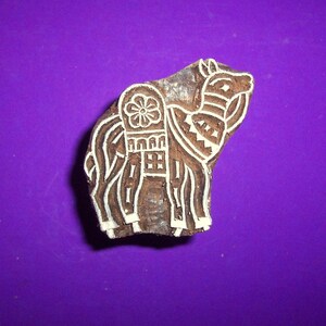May include: A wooden block printing stamp carved with a detailed design of a camel. The camel is decorated with white paint and has a floral pattern on its back.
