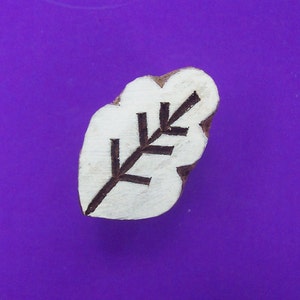 May include: A leaf-shaped wooden stamp with a white leaf design and dark brown veins. The stamp is set against a purple background. This stamp is ideal for crafting and scrapbooking projects.