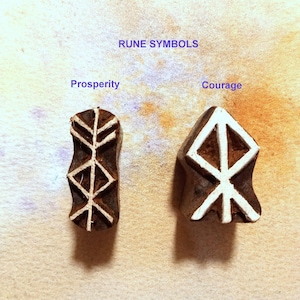 May include: Two wooden rune symbols, one for prosperity and one for courage. The symbols are carved in white on a dark brown background.