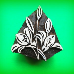 May include: A dark, triangular wooden block with a white floral design. The design features stylized lilies and buds, with a detailed outline. The block is set against a bright green background, highlighting the contrast between the dark wood and the white floral pattern.