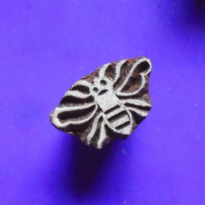 May include: A wooden block printing stamp featuring a white bee design. The bee has a striped body and wings. The stamp is brown and has a rough texture.