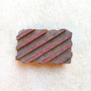 May include: A rectangular wooden block for textile printing featuring a series of raised, diagonal stripes across its surface. The wood has a natural brown tone with remnants of a reddish-brown pigment visible on the elevated striped sections.
