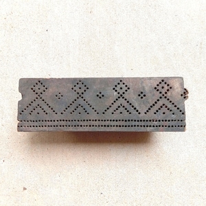 Könnte beinhalten: Ein rechteckiger, dunkelbrauner Holzblock für den Textildruck mit einem sich wiederholenden geometrischen Muster aus kleinen gebohrten Löchern. Das Design besteht aus einer horizontalen Punktreihe am unteren Rand und Rauten.