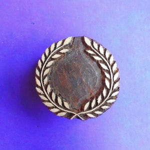 May include: A circular wooden stamp featuring a laurel wreath design. The central area is dark brown, surrounded by a light-coloured wreath. The background is a solid blue. This stamp is likely used for crafting or decorative purposes.