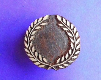 Puede incluir: Un sello de madera redondo con un diseño de corona de laurel. El sello tiene un centro marrón oscuro y una corona de color claro. El fondo es de color azul sólido. Este sello se utiliza probablemente para fines decorativos o artesanales.