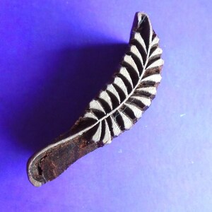 May include: A wooden block printing stamp with a carved design of a fern leaf. The stamp is brown and the leaf is white.