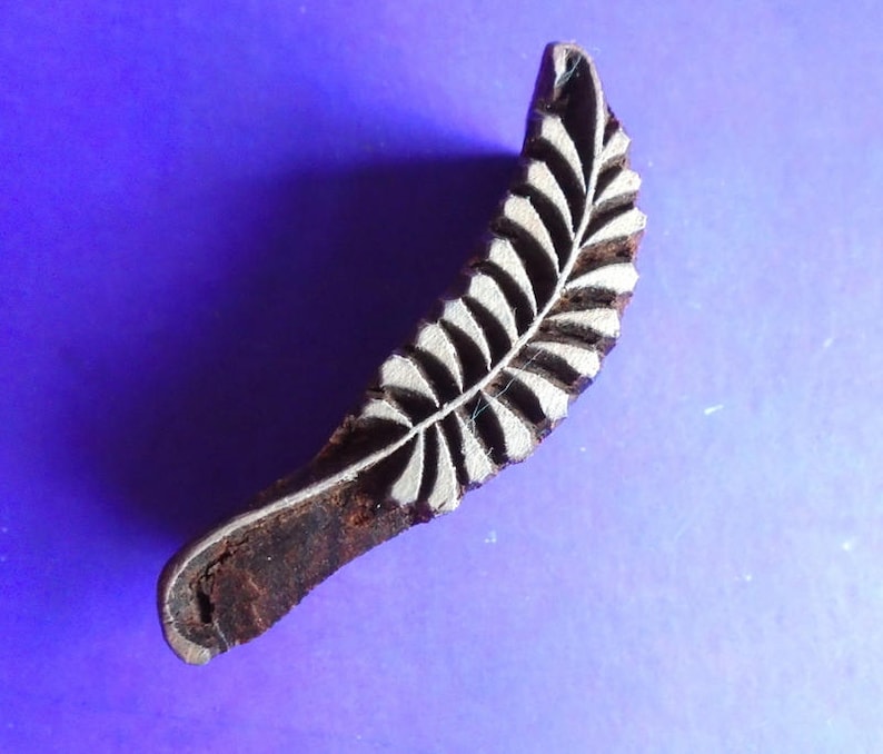 May include: A wooden block printing stamp with a carved design of a fern leaf. The stamp is brown and the leaf is white.