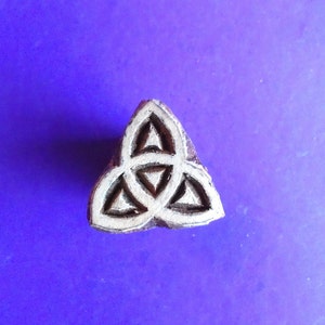 May include: A triangular, wooden stamp with a carved Celtic knot design. The stamp is a dark brown color and sits against a purple background. The knot is made up of three interlocking shapes.
