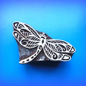 May include: A wooden block carving of a dragonfly with intricate details. The dragonfly is carved in white against a dark brown background.