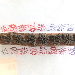 May include: A wooden block stamp with a floral design, used for printing. The stamp is dark brown with intricate carved details. Printed examples in red and blue ink show the floral pattern.