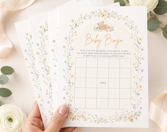 May include: A stack of white paper baby bingo game cards featuring a floral arch border in yellow, pink, and green. The cards include a central grid, a sleeping bear illustration, and the text Baby Bingo with instructions for the game.