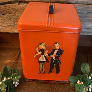 May include: An orange metal kitchen canister with a black handle and a decorative image of a dancing couple. The canister has a black base and is sitting on a wooden surface. Decorative greenery is placed on either side of the canister.