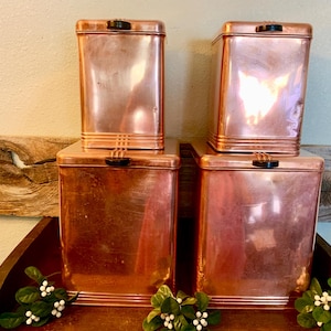 May include: Four copper-colored metal canisters with black handles. Two are stacked on top of the other two. The canisters have a rectangular shape with a slightly rounded top and bottom. The canisters are arranged on a wooden tray.