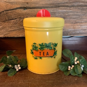May include: A vintage yellow tea canister with a red knob on the lid. The canister features a label with the word "TEA" in white letters, surrounded by green leaves. The canister is a classic kitchen storage item.