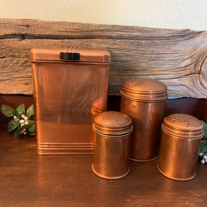 May include: A set of four copper-colored kitchen containers consisting of one large rectangular canister with a black handle on the lid and three smaller cylindrical shakers with perforated lids, all featuring a smooth, metallic finish.