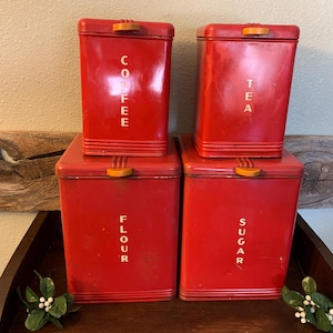 May include: Set of four vintage red metal kitchen canisters. The canisters are labeled "Coffee", "Tea", "Flour", and "Sugar" in yellow lettering. Each canister has a rectangular shape with a lid and a small wooden handle.