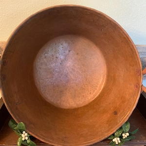 May include: A large, round, hammered copper basin with a wide opening and two circular side handles. The interior surface shows a textured, metallic finish with variations in color, while the exterior features a warm, reddish-brown patina.