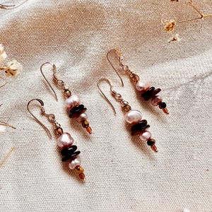 Könnte beinhalten: Vier Paar handgefertigte Ohrringe mit einem Anhänger-Design. Jeder Ohrring hat eine Kombination aus rosa Perlen, dunkelbraunen Steinen und kleinen goldfarbenen Perlen. Die Ohrringe werden auf einem neutralfarbenen Stoff präsentiert.