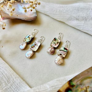May include: Two pairs of drop earrings featuring rectangular abalone shell pieces with iridescent green, purple, and blue tones. Each earring is accented with a single, irregular, pale pinkish-white baroque pearl suspended from a silver-toned hook.