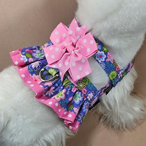 May include: A floral dog harness with a pink polka dot bow. The harness features a blue floral pattern with pink trim and a silver metal ring for leash attachment. The bow is a bright pink with white polka dots.