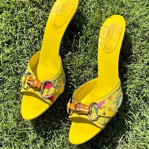 Peut inclure: Une paire de mules à bout ouvert jaune vif avec une tige en toile à motif floral, un détail mors de cheval en bambou sur le devant et un talon fin. La semelle intérieure jaune porte le nom de la marque Gucci embossé au centre.