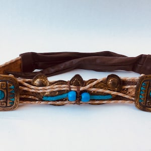Może przedstawiać: Brązowy skórzany pasek z tkanym wzorem i akcentami turkusowymi i złotymi. Pasek ma dwie duże kwadratowe klamry z turkusowymi kamieniami w środku.
