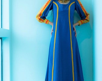 May include: A long, blue dress with yellow and orange trim. The dress features long sleeves with patterned cuffs and a matching trim at the hem. The dress is worn with white boots and a gold necklace.